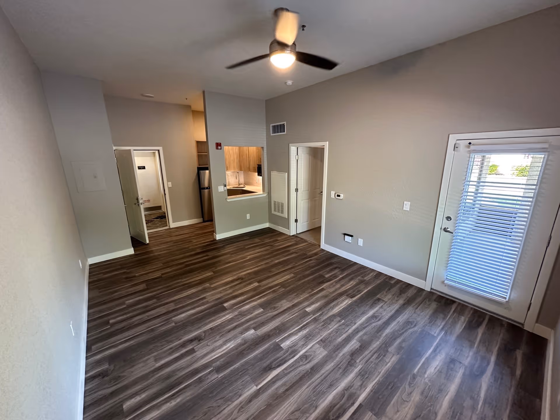 Empty room with wood flooring, beige walls, a ceiling fan with light, a small kitchen area with a stainless steel refrigerator and sink, two open doors, and a glass door with blinds leading outside.