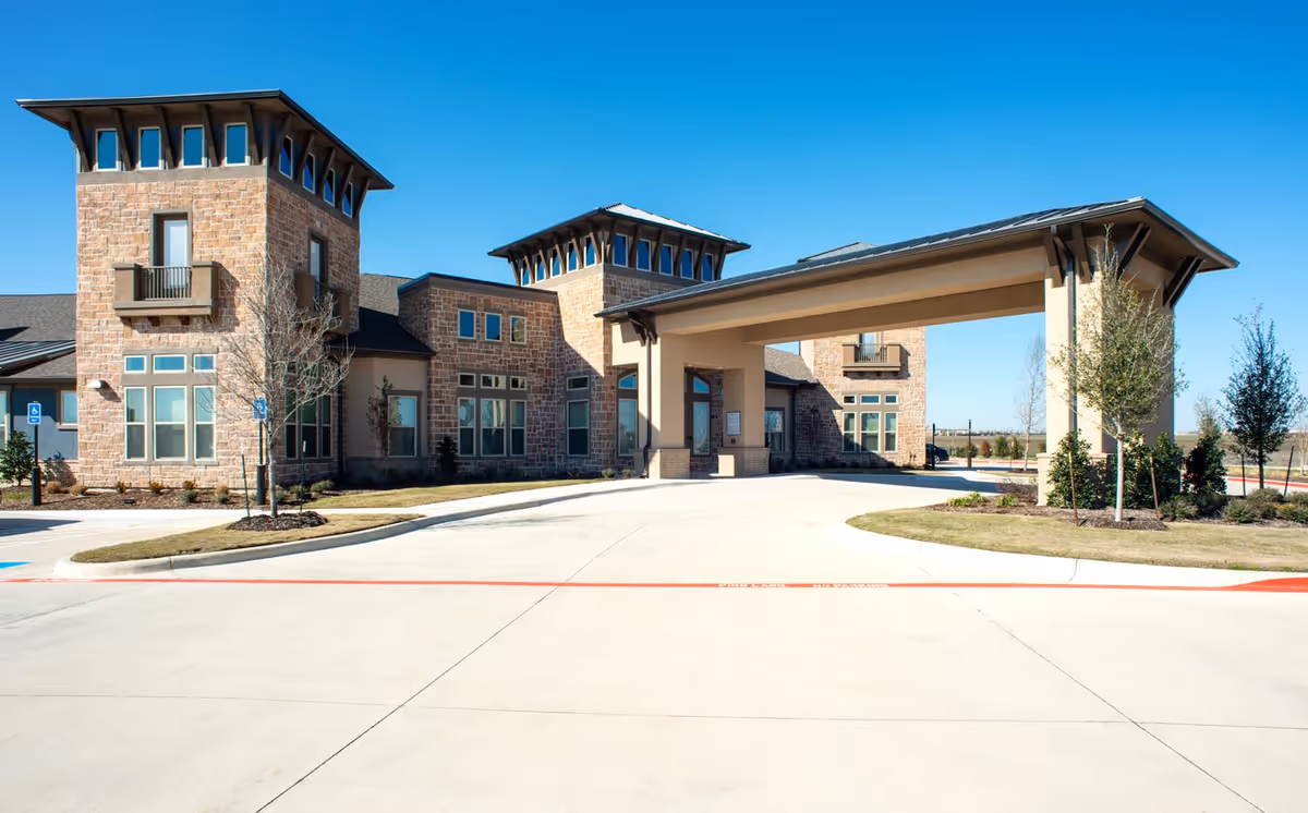 Front exterior view of Discovery Village at Alliance Town Center Assisted Living facility showing a large covered entrance with stone and beige walls, multiple windows, and a clear blue sky.