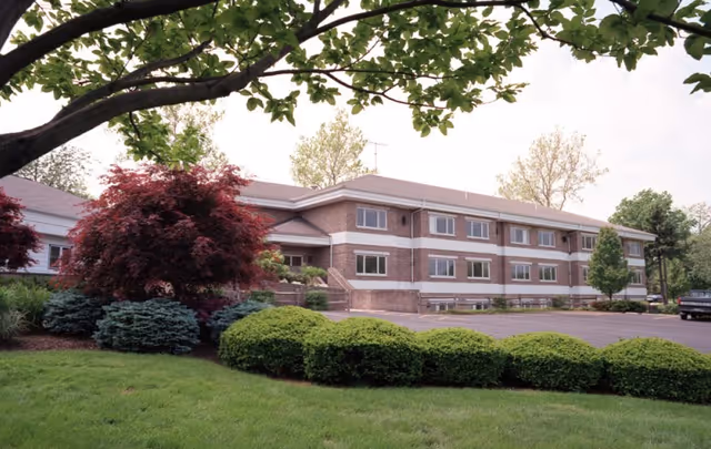 Exterior view of a two-story brick building with multiple windows, surrounded by well-maintained landscaping including green bushes, trees, and a grassy lawn. A paved parking area is visible to the right side of the building.