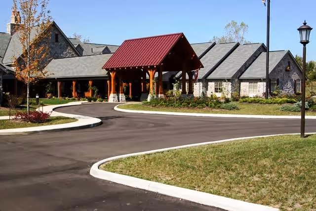 Exterior view of a senior living facility building with a stone facade and a red metal roof over the entrance. The driveway curves in front of the building, surrounded by landscaped grass and a few trees. A street lamp is visible on the right side.