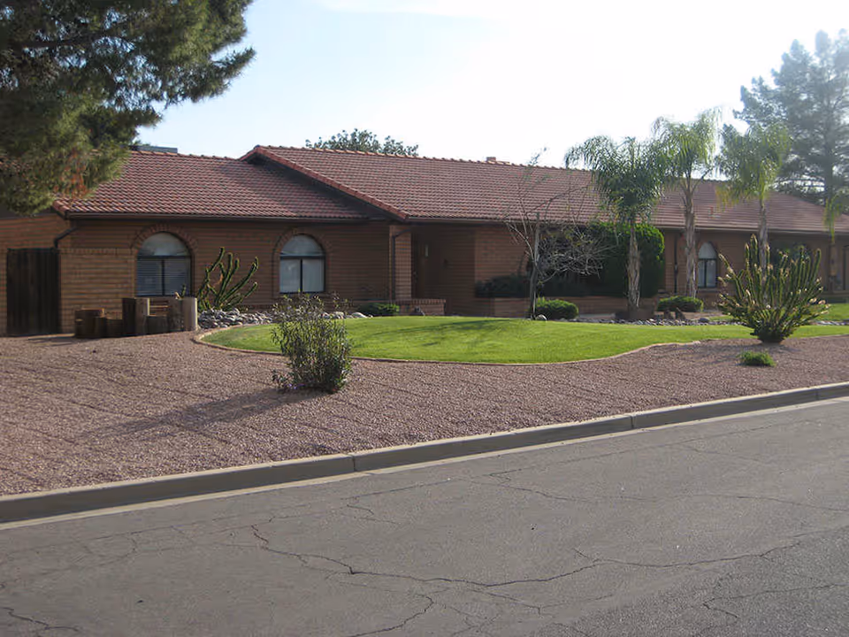 Single-story brick building with a red tiled roof, surrounded by a landscaped yard featuring green grass, palm trees, cacti, and other desert plants, viewed from across a paved street.