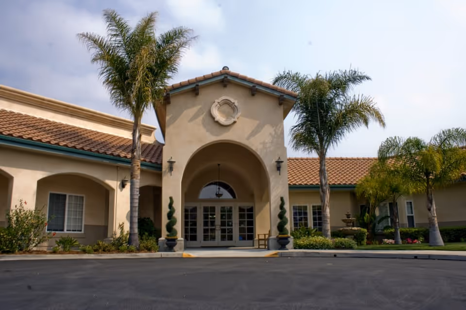 Front exterior view of The Village at Sydney Creek facility with a beige stucco building, arched entrance, tiled roof, palm trees, and landscaped greenery.