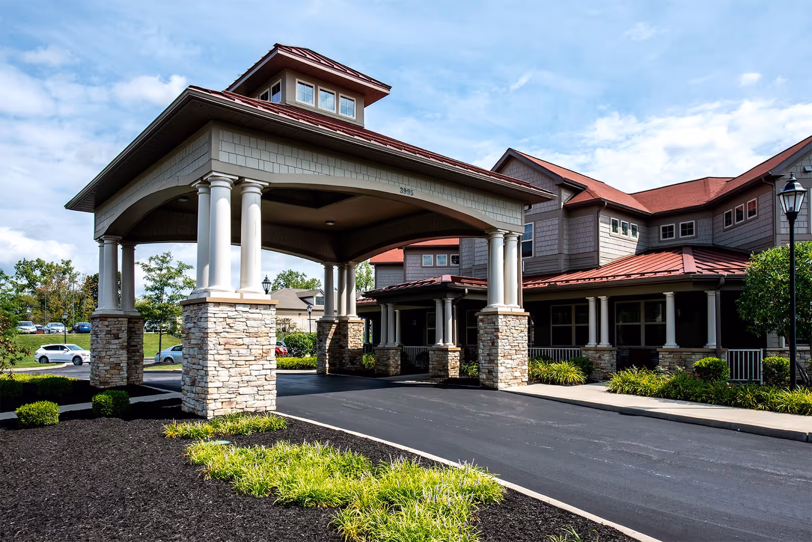 Exterior view of Beachwood Commons by New Perspective showing the entrance with a covered driveway supported by stone and white columns, landscaped greenery, and a multi-story building with red roofs under a partly cloudy sky.