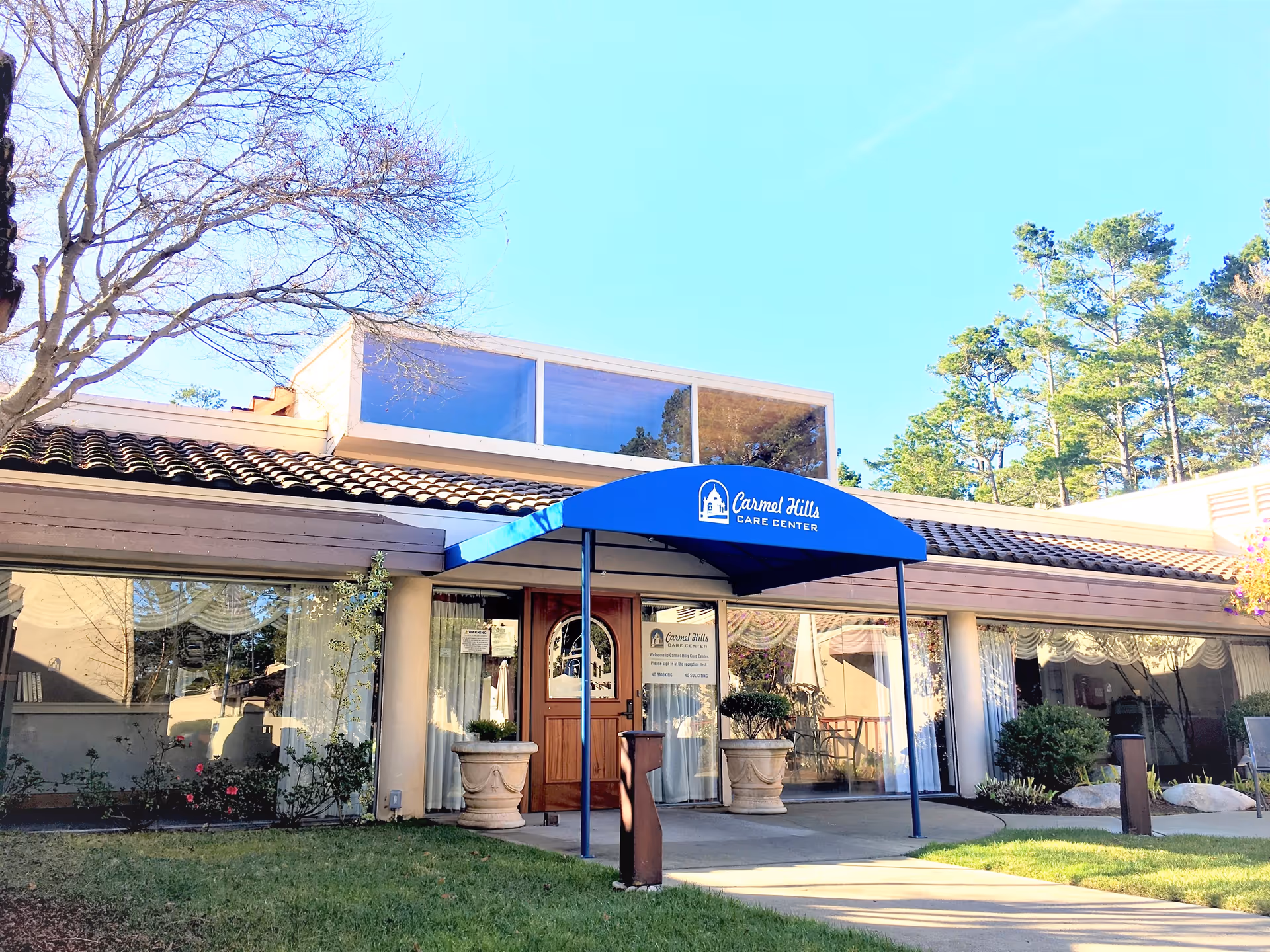 Exterior view of Carmel Hills Care Center building with a blue awning over the entrance. The building has large windows, a tiled roof, and is surrounded by trees and greenery.
