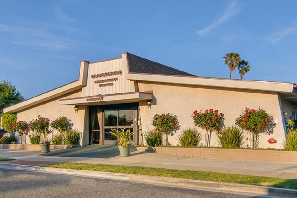 Exterior view of Orangegrove Rehabilitation Hospital building with a slanted roof, entrance doors, and landscaping including rose bushes and palm trees under a clear blue sky.
