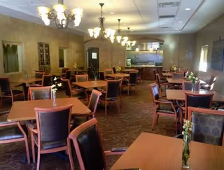 A dining room with multiple wooden tables and chairs arranged neatly. Each table has a small vase with flowers. The room has warm lighting from chandeliers hanging from the ceiling and a kitchen area visible in the background.