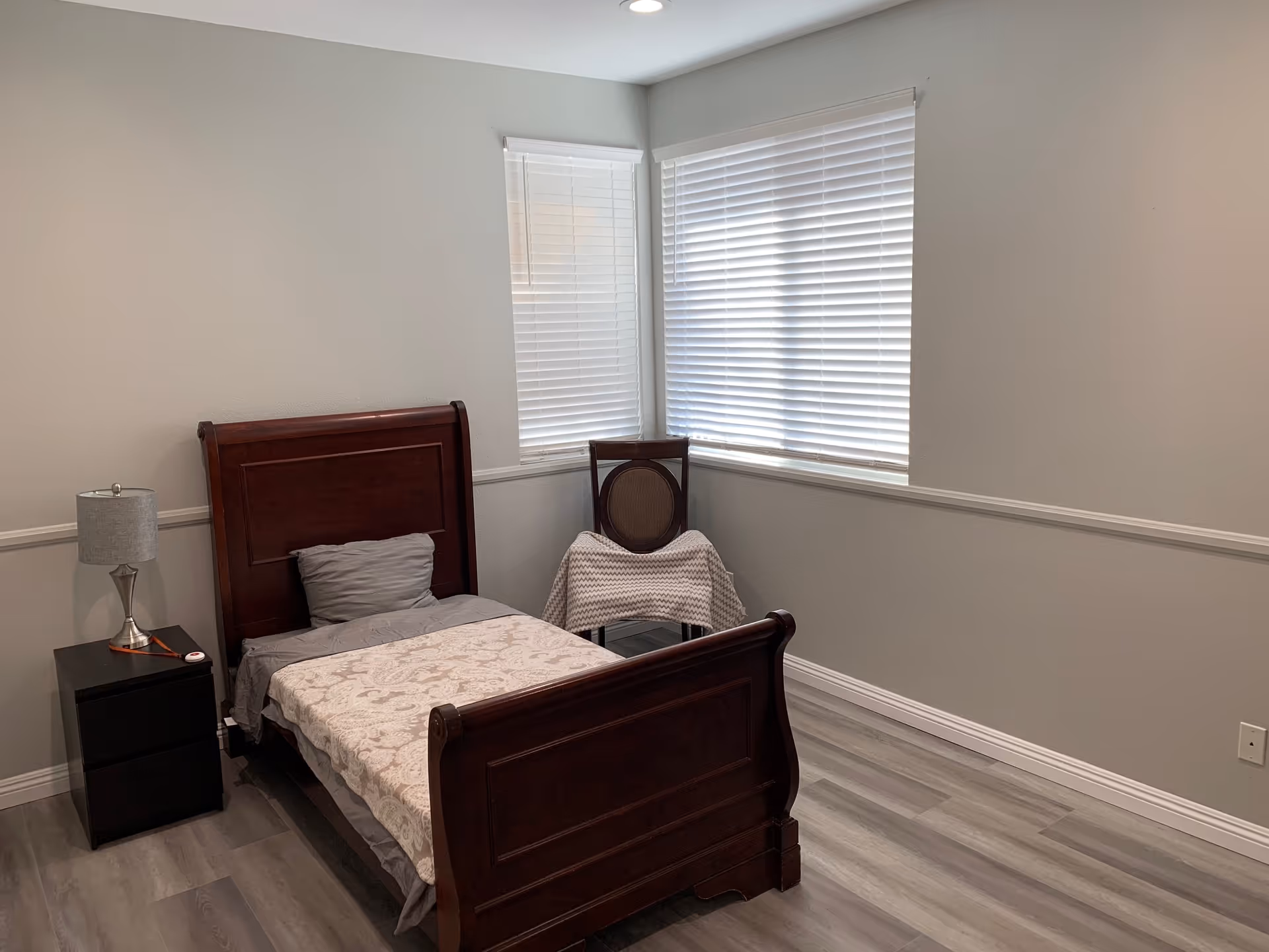 A simple bedroom with a single wooden bed with gray bedding and a patterned blanket. Next to the bed is a black nightstand with a silver lamp. There is a wooden chair with a blanket draped over it in the corner near two windows with white blinds. The walls are light gray and the floor has gray wood-like flooring.