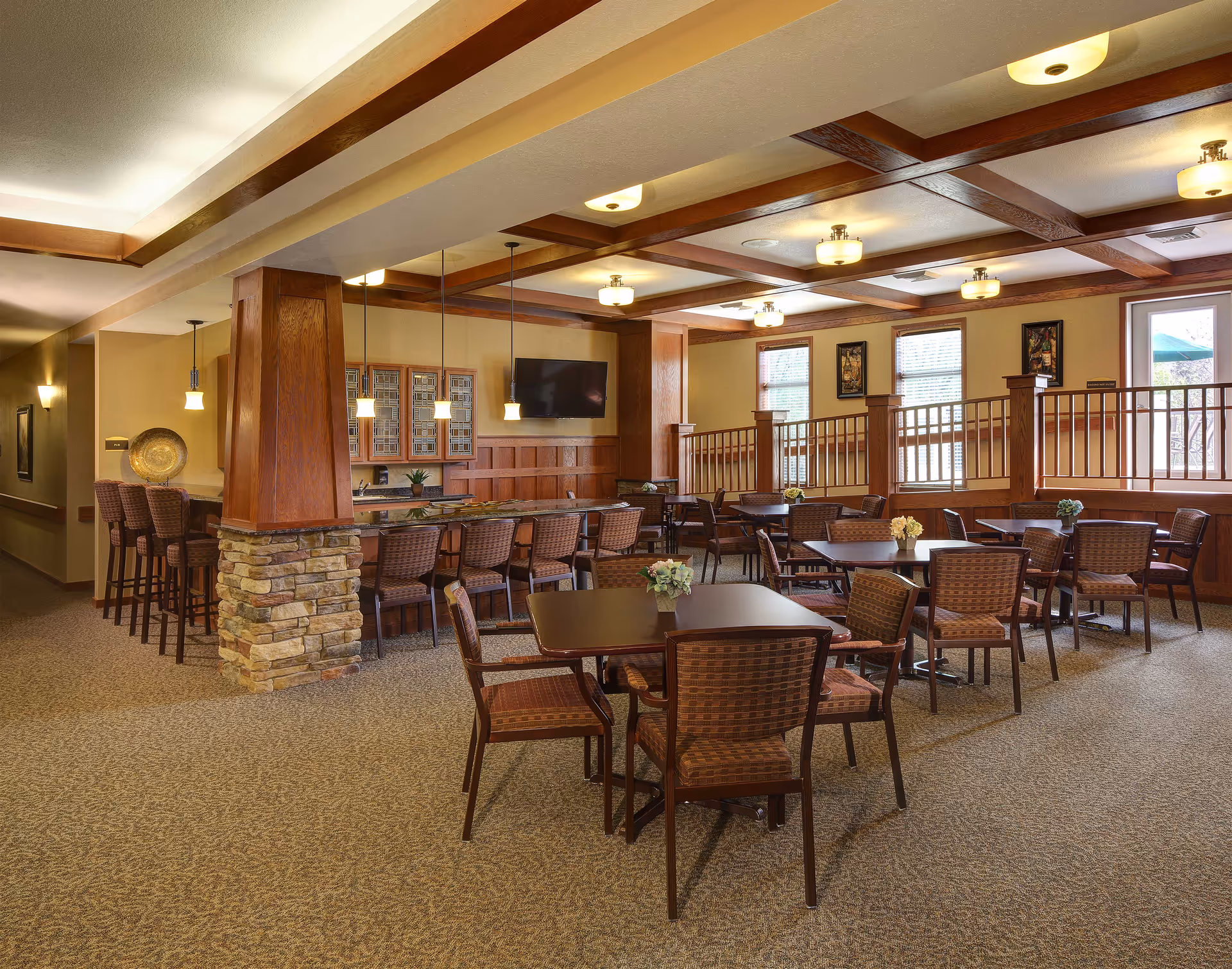 A spacious dining area in a senior living facility with multiple square tables and cushioned chairs arranged neatly. The room features warm wood accents, a stone pillar, pendant lights hanging over a bar counter with high chairs, and a wall-mounted TV. Large windows allow natural light to brighten the space.