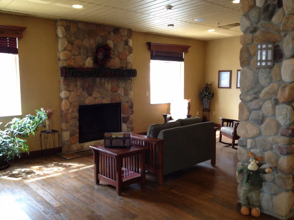 A cozy living room with a stone fireplace, wooden furniture including a sofa and chairs, a small table with a decorative box, potted plants, and two windows with wooden blinds letting in natural light. A stone pillar is visible on the right side with a small stuffed moose toy placed at its base.