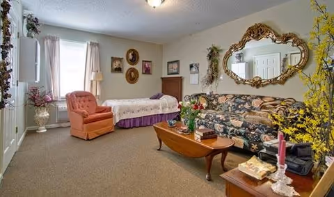 A cozy living space with a floral patterned sofa, a wooden coffee table with flowers and books, a coral upholstered armchair, and a bed with a white and purple bedspread. The room has beige walls decorated with framed pictures and a large ornate mirror. There are windows with sheer curtains letting in natural light.