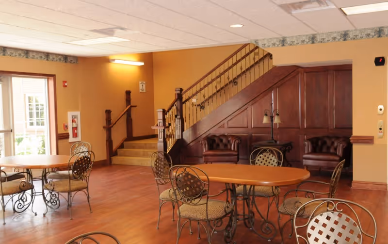 Interior view of a senior living facility common area with wooden floors, round tables with metal chairs, two leather armchairs under a staircase with wooden paneling, and a lamp on a small table. There is a staircase leading up and a door with windows letting in natural light.