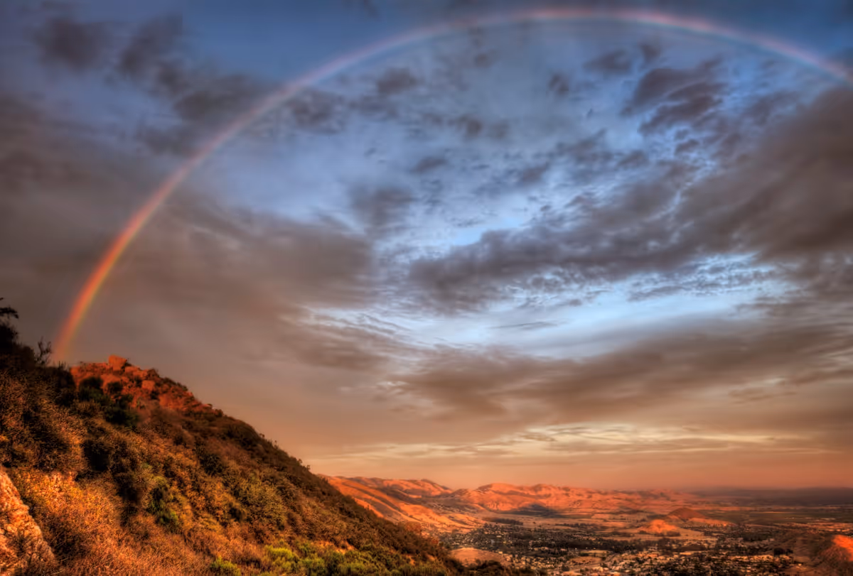A scenic landscape view of a hillside covered with bushes and trees under a cloudy sky with a rainbow arching over the hill. The distant valley and hills are bathed in warm sunlight.