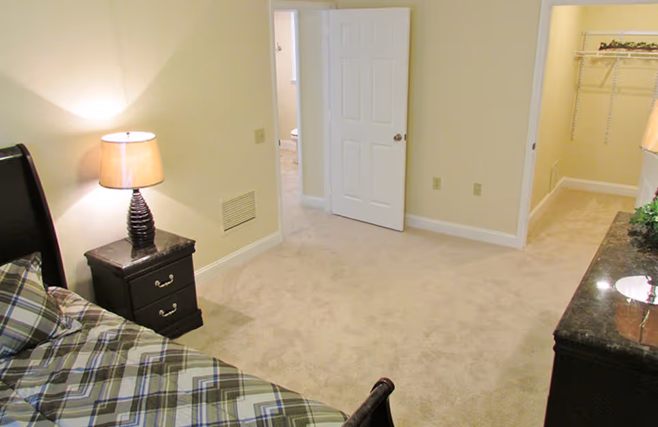 A bedroom with beige carpet and walls, featuring a bed with a plaid patterned bedspread, a dark wooden nightstand with a lamp, an open door leading to a bathroom, and an open walk-in closet with wire shelving.
