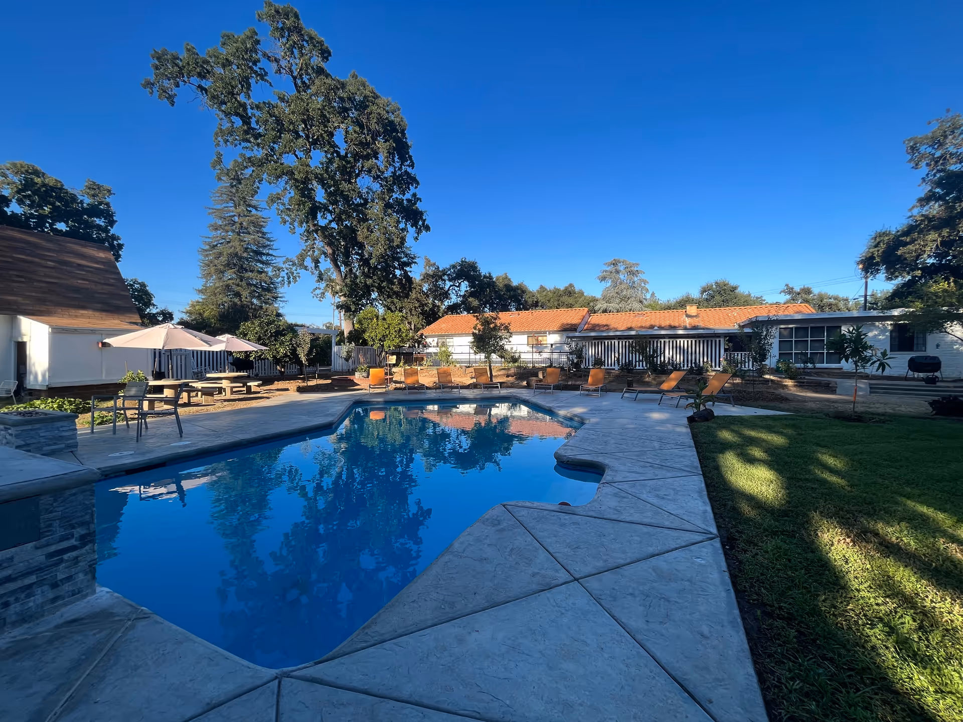Outdoor area of a senior living facility featuring a swimming pool surrounded by a concrete deck, lounge chairs, picnic tables with umbrellas, and trees under a clear blue sky.