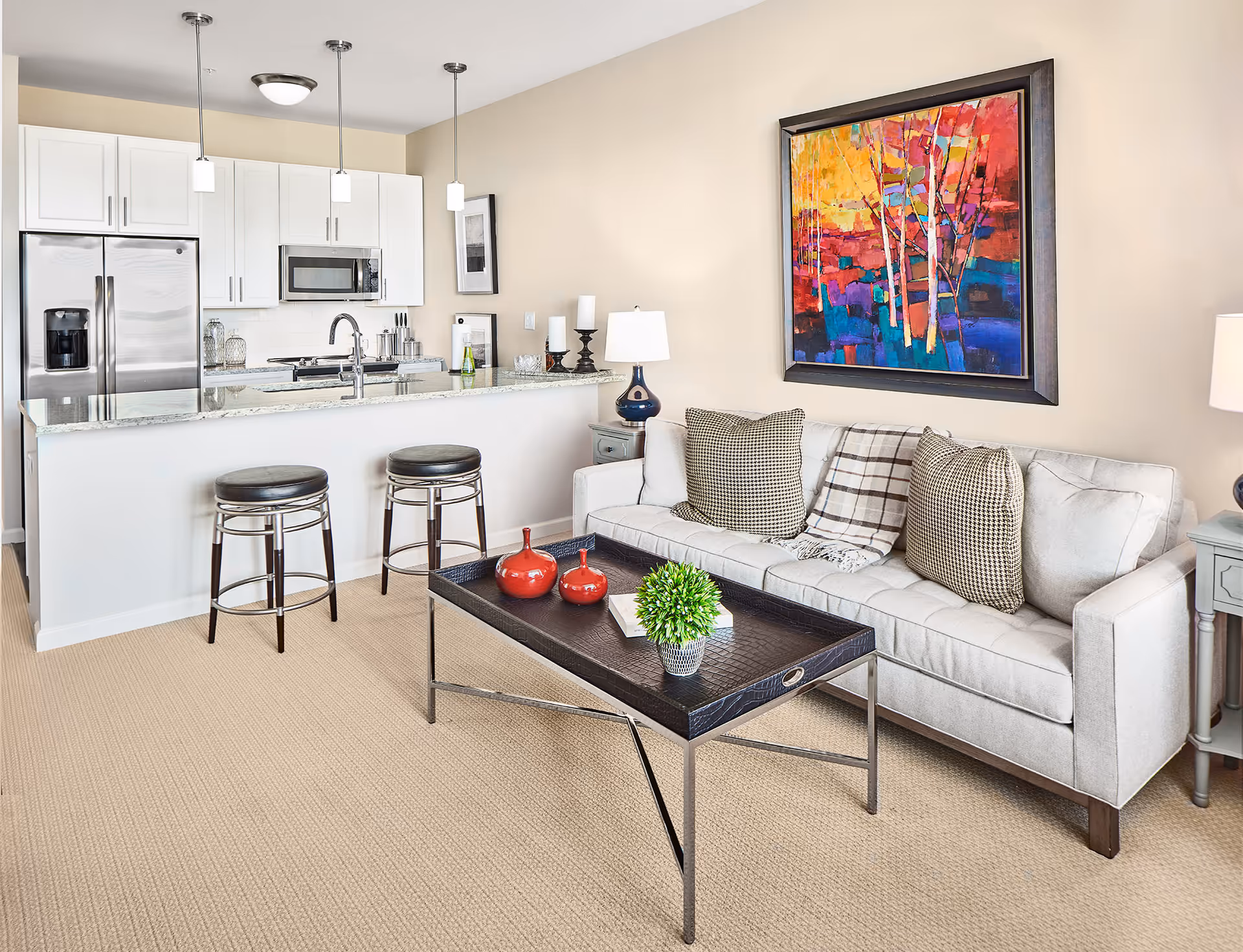 A modern living room and kitchen area featuring a light beige sofa with patterned pillows and a plaid throw, a black metal and leather coffee table with decorative items including two red vases and a small green plant, and a kitchen with white cabinets, stainless steel refrigerator, microwave, and a granite countertop with two black bar stools. A colorful abstract painting hangs on the wall above the sofa.