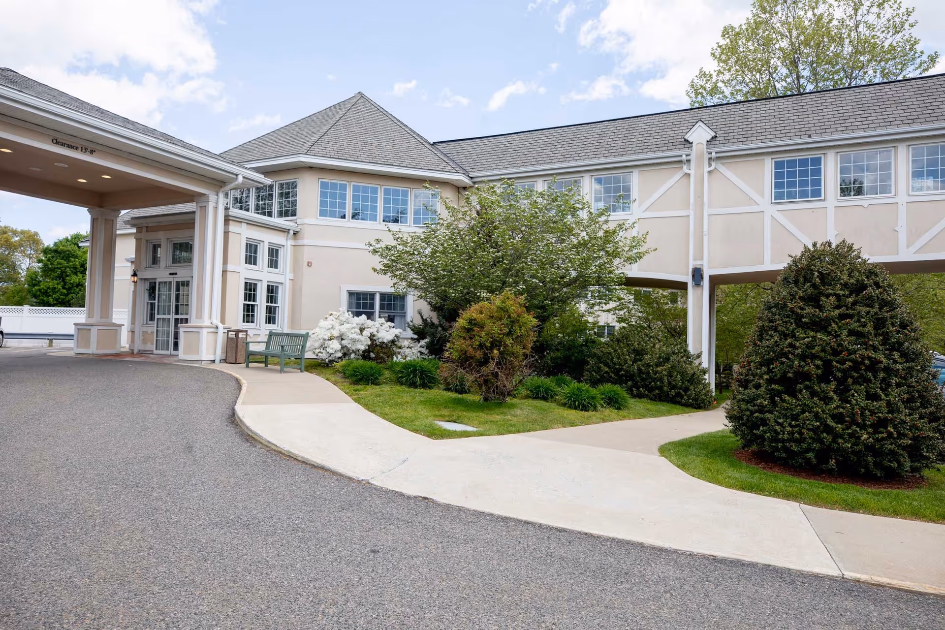 Exterior view of Edelweiss Village senior living facility showing a covered entrance with a clearance sign, a pathway, green bushes, trees, and a beige building with multiple windows under a partly cloudy sky.